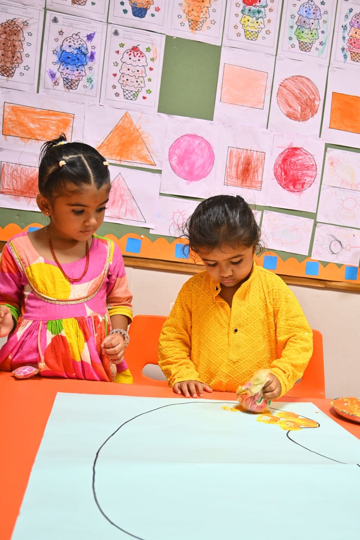 Two young students painting on a large sheet of paper below a wall of their artwork