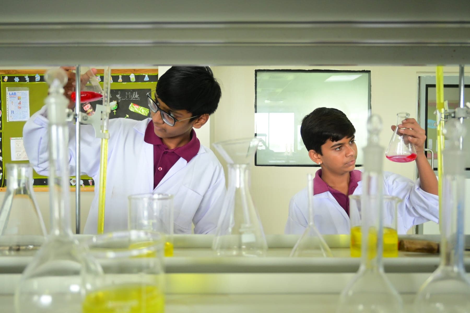 Two students in lab coats measuring liquids into flasks in the school chemistry lab